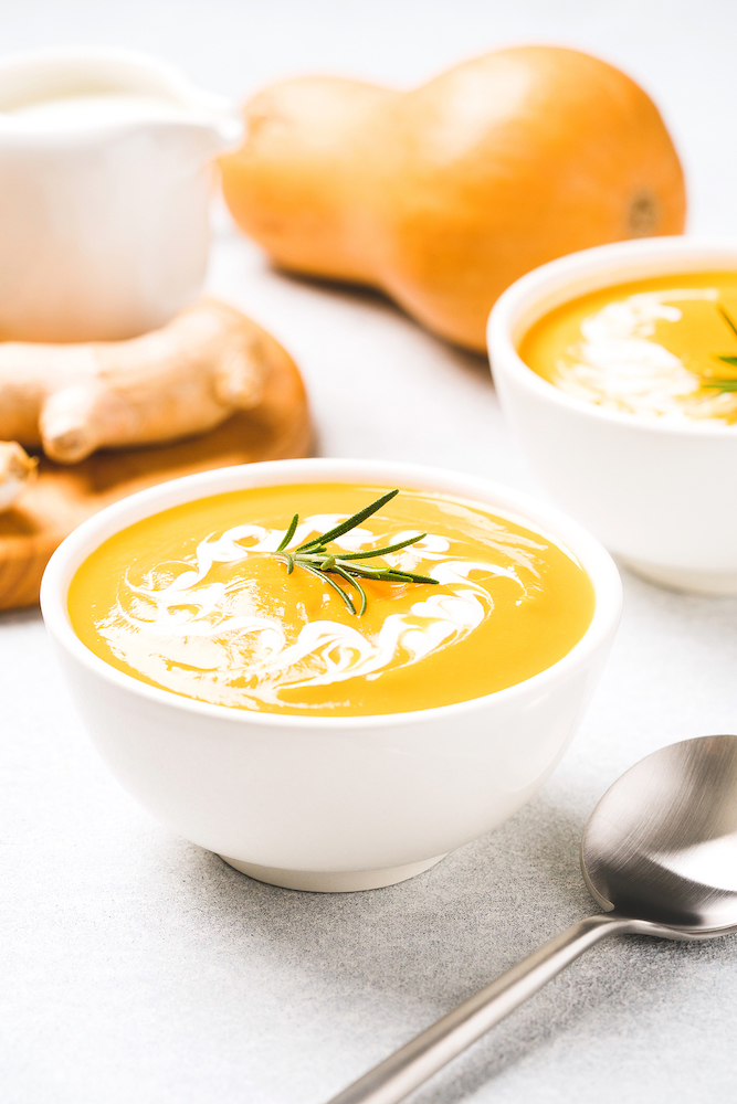 White bowl with thick butternut squash bisque garnished with heavy cream and rosemary on gray concrete table. Bowl of pumpkin cream soup with ginger. Vertical image, minimal style