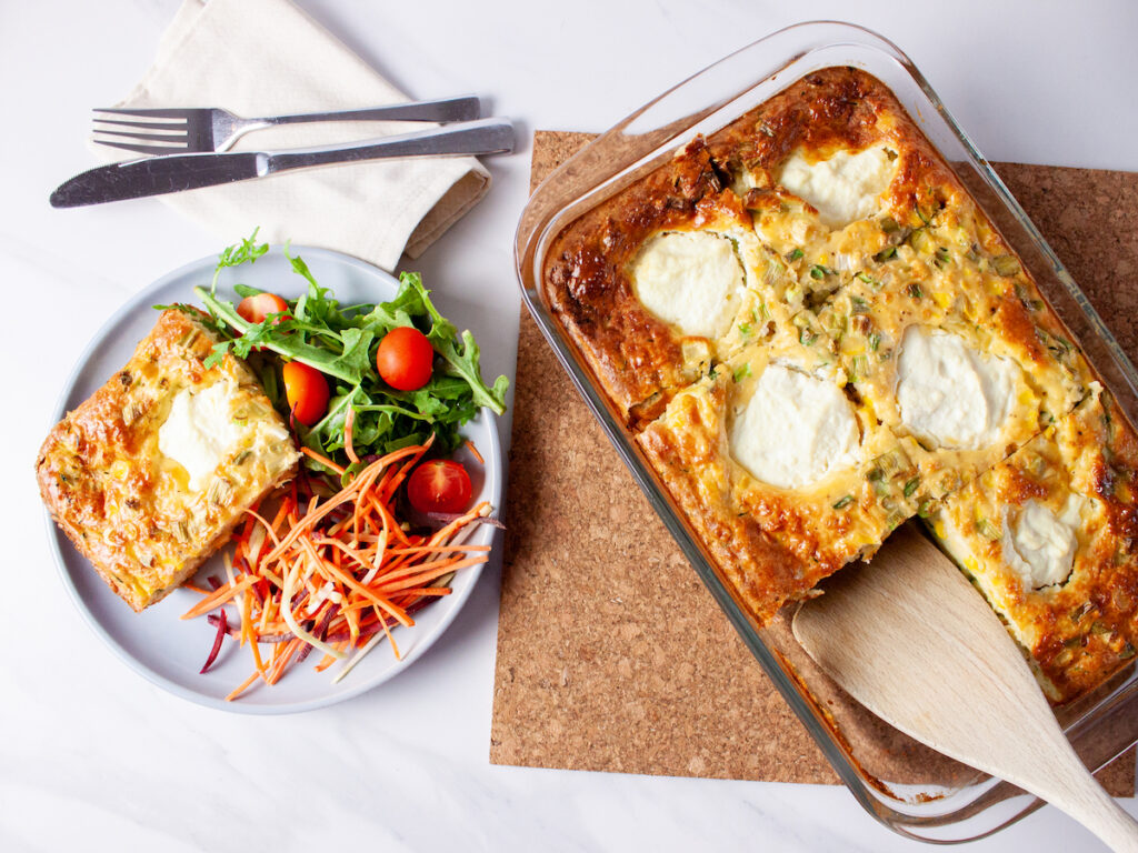 Sliced Zucchini and Ricotta Bake in a clear baking tray next to a plate a slice of Zucchini and Ricotta Bake with salad