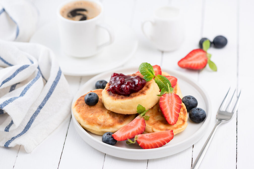 Cottage cheese pancakes or Syrniki with strawberries, blueberries, berry jam and cup of black coffee espresso. Tasty breakfast on white table